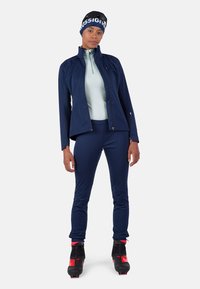 Marineblaues Sportjackett mit hohem Kragen über einer hellblauen Basisschicht, kombiniert mit marinen Hosen und schwarzen Skischuhen mit roten Akzenten.