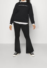 Maglione nero di Calvin Klein con logo bianco, indossato sopra una camicia bianca con colletto. I pantaloni neri a zampa d'elefante e le sneakers nere completano l'outfit.