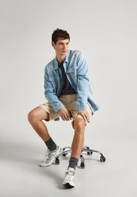 Giacca di jeans azzurro chiaro su una maglietta scura, pantaloncini beige, calze grigie e sneakers bianche. Seduto su una sedia da ufficio, gambe incrociate in modo casual.