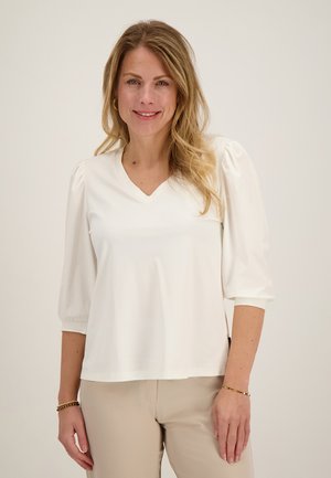 Vrouw met lang blond haar, die een witte V-hals blouse met opgeblazen mouwen en beige broek draagt tegen een neutrale achtergrond.