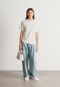 Λευκό κοντομάνικο μπλουζάκι με την επιγραφή "BOSS", σε συνδυασμό με ανοιχτόχρωμο τζιν παντελόνι με φαρδιές φάσες, λευκά αθλητικά παπούτσια και μικρή μπεζ τσάντα χειρός.