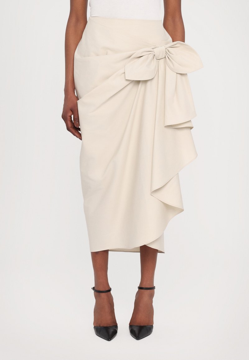 Jupe beige longueur genoux avec un grand accent en nœud sur le côté, présentant un drapé superposé et une texture lisse. Accompagnée de talons noirs.