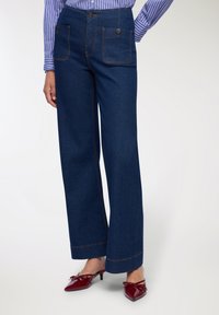 Pantaloni in denim blu scuro a gamba larga con due tasche frontali e cuciture arancioni a contrasto. Abbinati a mule rosse con punta.