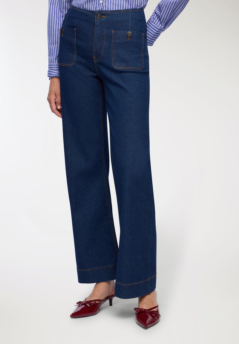 Pantaloni in denim blu scuro a gamba larga con due tasche frontali e cuciture arancioni a contrasto. Abbinati a mule rosse con punta.