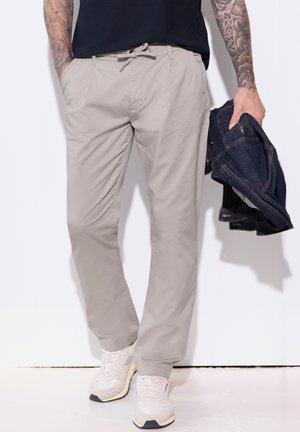 Homme portant un pantalon beige à cordon de serrage et des baskets blanches, tenant une veste en jean foncée pliée dans une main, avec les bras tatoués visibles.