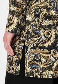 Tessuto nero con motivo paisley in oro e bianco; presenta maniche lunghe, aperture laterali e una texture liscia; include dettagli decorativi.