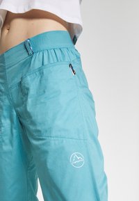 Light blue hiking pants in durable fabric with elastic waistband, side pocket, and a logo on the lower leg. Smooth texture and relaxed fit.