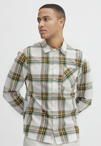 Camisa de manga larga de cuadros en beige, verde, amarillo y marrón. Presenta un cuello de botones, un bolsillo frontal y un dobladillo redondeado.