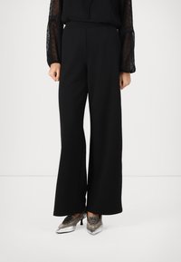 Femme portant un pantalon large noir, un blouse noire transparente avec des manches ballons et des escarpins argentés métalliques à bout pointu.