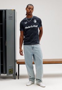 Camisa desportiva azul escura com padrão ondulado e logótipo branco, combinada com jeans largos azul claro e ténis brancos. Balcão e banco ao fundo.