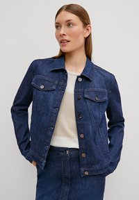 Veste en denim foncé avec quincaillerie argentée, deux poches poitrine, fermeture à boutons et design ajusté, associée à un haut clair et une jupe en denim assortie.