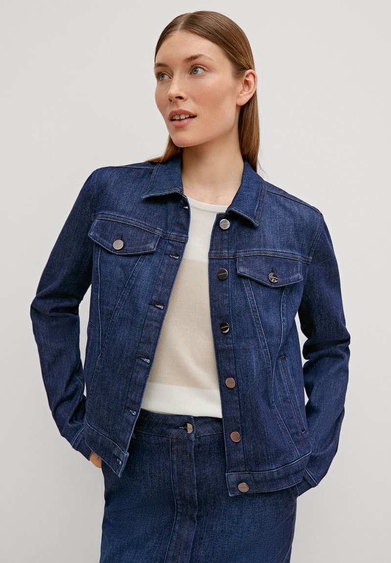 Veste en denim foncé avec quincaillerie argentée, deux poches poitrine, fermeture à boutons et design ajusté, associée à un haut clair et une jupe en denim assortie.