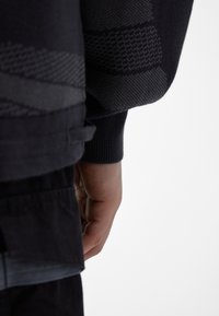 La main d'une personne repose le long de son corps, portant un pantalon sombre et un pull sombre à motifs, sur un fond clair et uni.