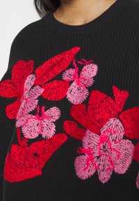 Svart stickad tröja med livfulla röda och rosa blommönster och en texturerad yta. Crew neck-design, närbild av blommornas detaljer.