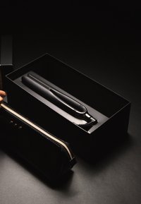 Prostownica do włosów w czarnym, matowym opakowaniu, obok teksturowanego czarnego etui z złotym suwakiem i ćwiekami. Tło jest ciemne.