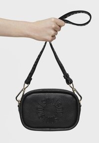 Czarna skórzana torba na ramię w kształcie owalu z wytłoczonym logo, z odpinanym paskiem z powtarzającym się wzorem tekstu. Złote akcenty.