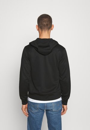 Personne aux cheveux courts de dos portant un sweat à capuche noir par-dessus une chemise blanche et un jean bleu sur un fond uni.