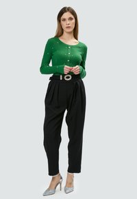 Maglietta verde a coste con maniche lunghe e perline decorative, abbinata a pantaloni neri a vita alta e scarpe con tacco argentate lucide.