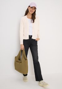 Lichtroze cardigan over een witte top, zwarte broek, met een groene puffer tas. Lichtpaarse pet en lichtgekleurde schoenen. Zachte texturen.
