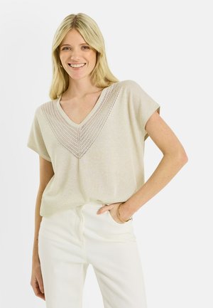 Mujer rubia sonriente con una blusa de punto beige de manga corta con escote en V y pantalones blancos, con una mano en el bolsillo, sobre un fondo blanco.