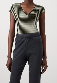 Top a V con righe in tessuto verde oliva scuro e nero, con maniche a cappuccio e un piccolo dettaglio del logo; abbinato a pantaloni scuri a costine con vita elastica.