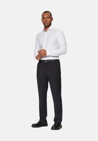 Camisa blanca de botón con una textura sutil, combinada con pantalones oscuros a medida y zapatos de vestir negros. El atuendo presenta un diseño ajustado.