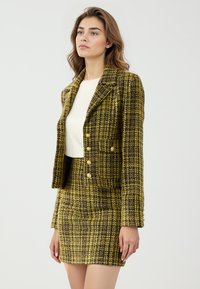 Ensemble blazer et jupe en tweed de couleur jaune, noir et blanc. Le blazer est orné de boutons dorés et a une coupe ajustée. Texture lisse et structurée.