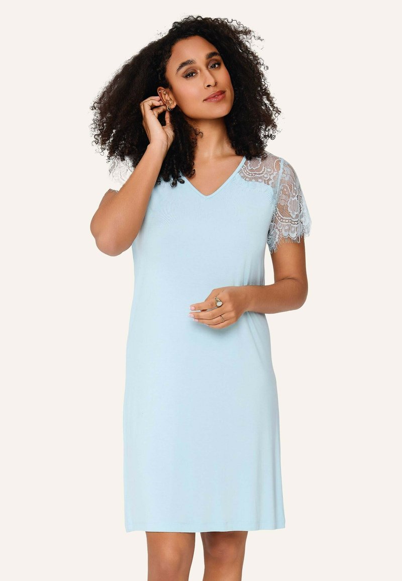 Frau mit lockigem dunklem Haar, die ein hellblaues knielanges Kleid mit kurzen Spitzenärmeln trägt, sie berührt ihr Ohr und hält ihre Hand in der Nähe der Taille.