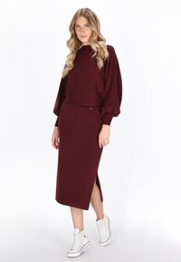 Bordeaux gebreide trui met brede mouwen, gecombineerd met een bijpassende midi-rok met een zijsplit. Witte hoge sneakers maken de look af.