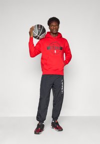 Rød hettegenser med "BULLS" logo, svarte bukser med sidebranding, og svarte/røde joggesko. Holder en basketball. Jevn tekstur.