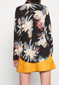 Langærmet sort skjorte med blomstermotiv i blå, creme og rød, med en buet kant og knapmanchetter. Bæres med en gul ruskindsnederdel.