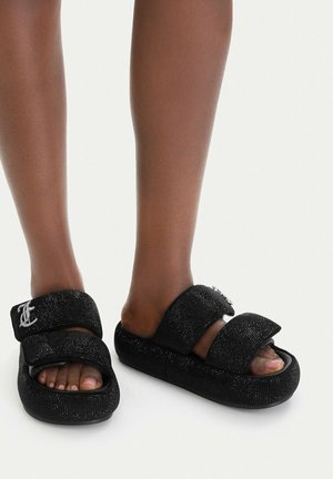 Barfuß mit schwarzen Plateau-Slide-Sandalen mit zwei funkelnden Riemen und einem silbernen dekorativen Logo auf einem Riemen.
