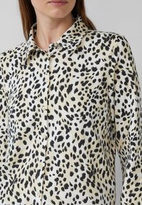 Chemise à imprimé léopard avec un fond crème et de grandes taches noires. Conçue avec un col et une fermeture à boutons, fabriquée en tissu léger.