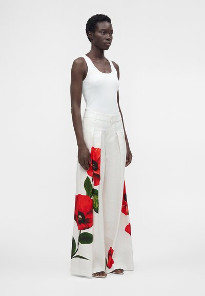 Femme debout portant un haut blanc sans manches et un pantalon blanc à jambes larges avec de grandes impressions de fleurs rouges et des feuilles vertes.