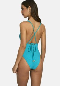 Un costume de bain une pièce turquoise avec une jambe haute, des bretelles croisées et un détail de dos cintré avec des liens ajustables et des accents dorés.