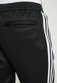 Černé sportovní kalhoty z texturované látky, s bílými svislými pruhy po stranách a malou kapsou na zádech.