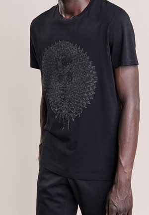 Homme portant un t-shirt noir à manches courtes avec un design géométrique abstrait argenté détaillé sur le devant, associé à un pantalon noir.
