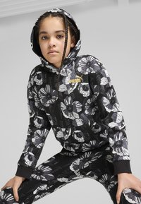Svart hoodie och leggings-set med ett blommönster i grått och vitt, med guldlogotypen som accent på bröstet och kontrasterande ärmslut.