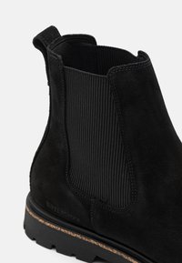 Bottine cheville en suede noir avec des panneaux latéraux élastiques, une finition texturée et une semelle en caoutchouc avec un accent en liège visible.