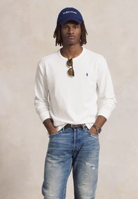 Giovane con dreadlocks che indossa un cappellino blu navy, una camicia bianca a maniche lunghe, occhiali da sole sul colletto, jeans strappati e un orologio al polso, con le mani in tasca.