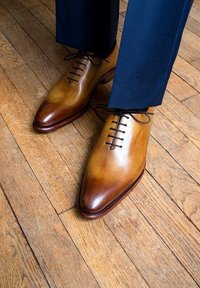 Chaussures en cuir couleur tan dégradé, avec des bouts pointus, des lacets noirs et une finition brillante, associées à un pantalon bleu marine sur un sol en bois.
