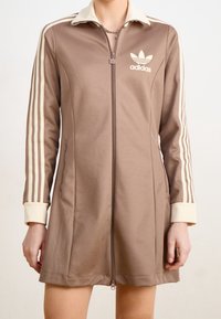 Braunes Reißverschlusskleid mit einem cremefarbenen Kragen und Manschetten, ausgestattet mit drei cremefarbenen Streifen an jedem Ärmel und einem Adidas-Logo auf der Brust.