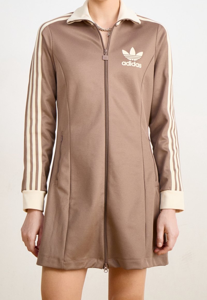 Braunes Reißverschlusskleid mit einem cremefarbenen Kragen und Manschetten, ausgestattet mit drei cremefarbenen Streifen an jedem Ärmel und einem Adidas-Logo auf der Brust.
