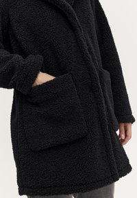 Zwarte fleece jas met een gestructureerd oppervlak, een brede kraag, voorzakken en een losse pasvorm. De manchetten en zoom zijn ook afgewerkt met een zachte afwerking.