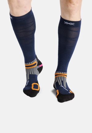 Beine mit marineblauen Kompressionssocken mit orangefarbenen, pinken und grauen Akzenten, stehend auf weißem Hintergrund.