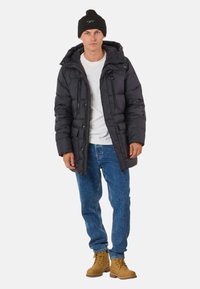 Black puffer jacket with a hood, zippered pockets, and ribbed cuffs, paired with blue jeans and tan work boots.