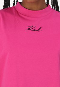 Rosa t-skjorte med høy krage og svart brodert "Karl"-logo. Glatt bomullsstoff, avslappet passform og minimalt design.