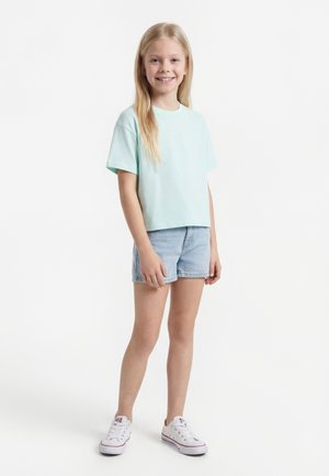 Fille souriante aux longs cheveux blonds portant un t-shirt bleu clair à manches courtes, un short en jean et des baskets blanches, debout devant un fond blanc.