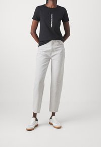 Osoba w czarnej koszulce Armani Exchange, jasnoszarych spodni wysoko w talii i białych trampkach z gumową podeszwą na białym tle.