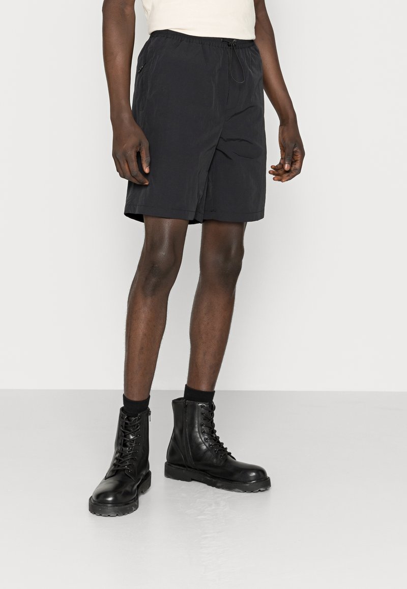 Homme portant un short noir avec cordon de serrage, des bottes de combat noires à lacets et des chaussettes noires sur un fond clair uni.